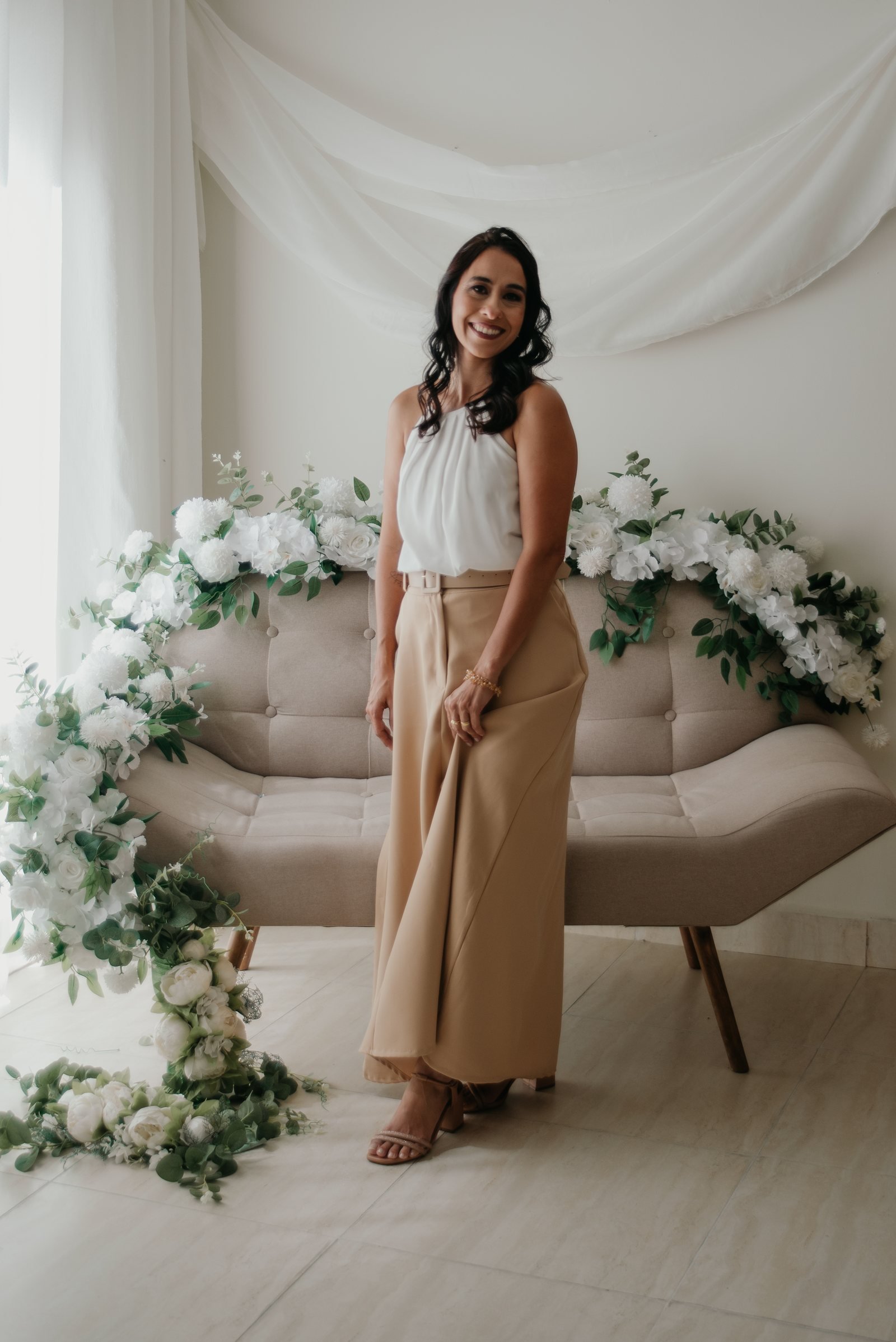 Fundadora da Florada de Ipê em pé, ao lado de um sofá bege decorado com flores brancas